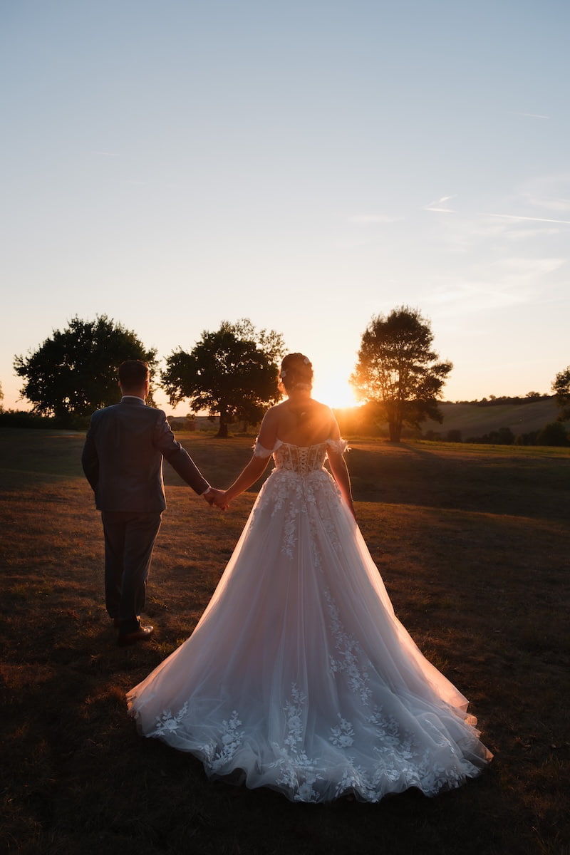 Mariés de dos se tenant la main face au coucher du soleil dans une lumière douce