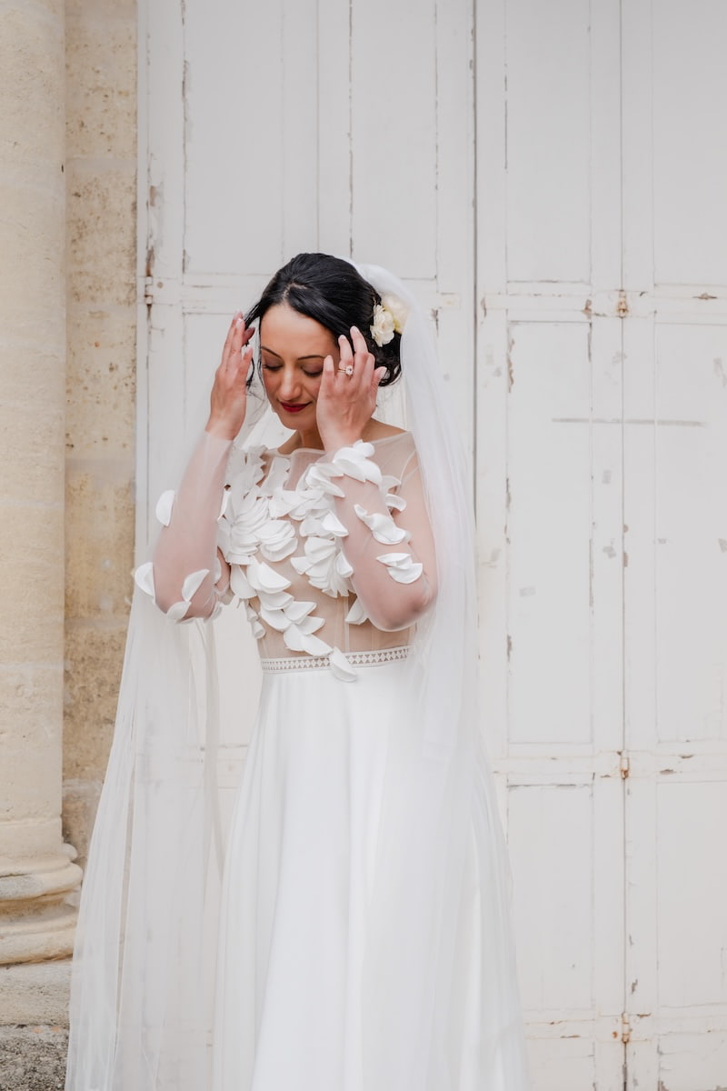 Mariée de face se tenant les cheveux devant une grande porte blanche d'une église en Île-de-France